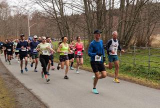 A group of people run on an outdoor trail in a wooded area, in a close group during a race at Royal Roads University